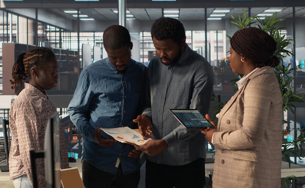 Four colleagues standing in an open-plan office reviewing printed performance charts and a tablet.